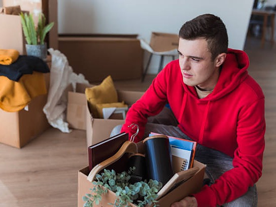 A student crouching beside packed cardboard boxes in an empty apartment, holding a box of books and plants